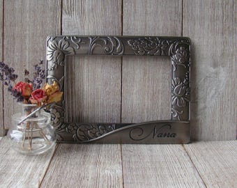 May include: Silver picture frame with an ornate floral design and the word "Nana" engraved on the bottom. The frame is sitting on a wooden surface.