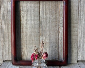 May include: A rectangular dark reddish-brown wooden frame with rounded corners positioned behind a small, clear glass jar containing a bouquet of dried pink roses and sprigs of lavender with a small metal handle attached to the rim.