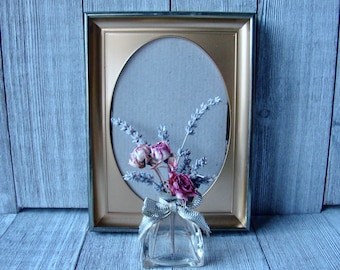 May include: A gold-framed oval picture frame with a dried flower arrangement in a clear glass vase with a gold bow.