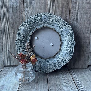 May include: A decorative, light blue ceramic picture frame with a floral and lace design. The frame has a central opening and a decorative border. A small glass vase with dried flowers sits in front of the frame.