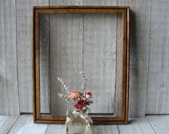 May include: A rectangular wooden picture frame with a dark brown burl wood grain finish. In front of the frame sits a small, clear glass bottle containing a bouquet of dried lavender sprigs and small, faded pink rose blossoms.