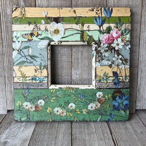 May include: A square wooden picture frame featuring a distressed, multi-colored horizontal striped background in shades of yellow, green, and blue. The frame is decorated with detailed botanical illustrations of white, pink, and blue flowers and a butterfly.