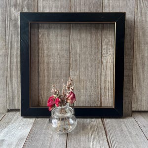 May include: A square black picture frame with a thin gold inner border, positioned behind a small, clear glass jar containing a bouquet of dried pink roses and sprigs of lavender. The frame has a smooth finish and a simple, clean design.