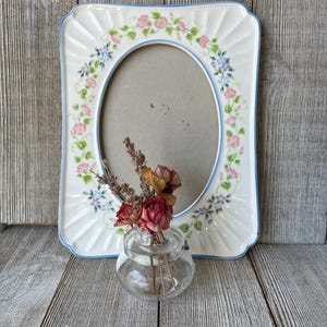 May include: A rectangular ceramic picture frame with a central oval opening, featuring a white base with blue trim and a floral pattern of pink and blue flowers with green vines. A small clear glass jar holds dried red roses and lavender stems.