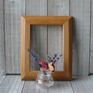 May include: A wooden picture frame with a natural wood finish. The frame is empty and is sitting on a wood surface. A small glass vase with dried flowers is in front of the frame.