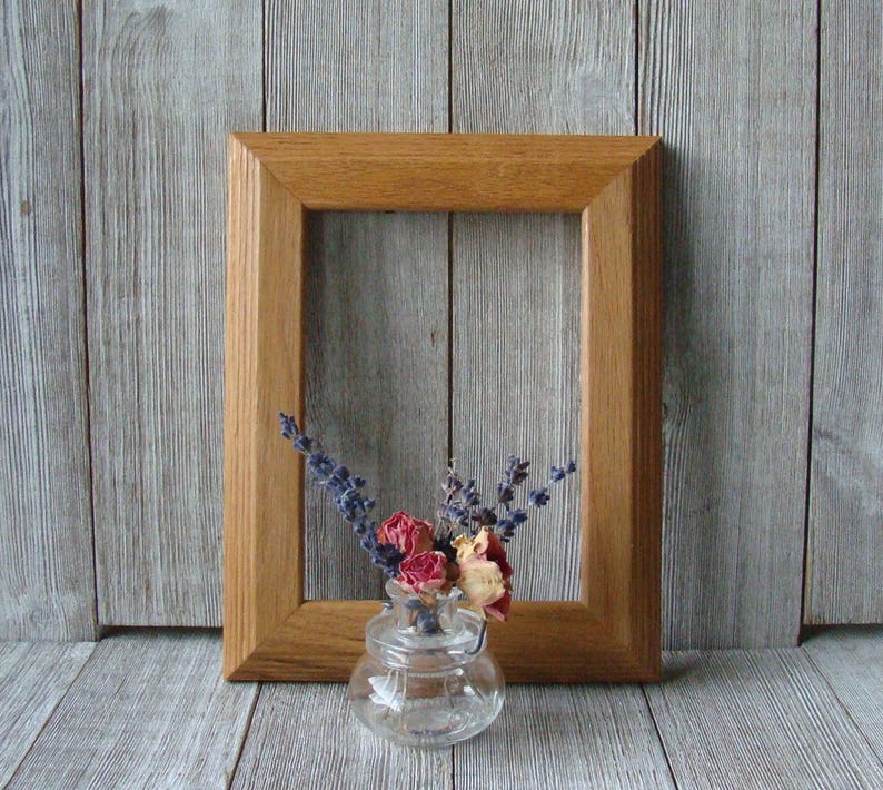 May include: A wooden picture frame with a natural wood finish. The frame is empty and is sitting on a wood surface. A small glass vase with dried flowers is in front of the frame.