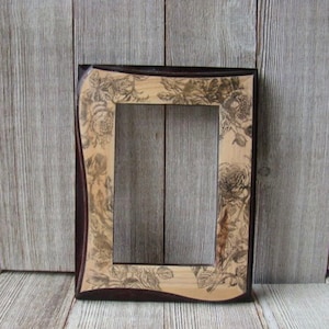 May include: A dark brown wood picture frame with a decorative floral design etched into the wood. The frame has a slightly wavy edge.