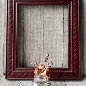 May include: A burgundy-colored picture frame with a textured pattern on the border. The frame is empty, with a small glass jar containing dried flowers in front of it. The background is a wooden surface.