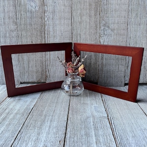 May include: Two rectangular, dark red wooden picture frames are displayed on a weathered, gray wooden surface. A small, clear glass vase with dried flowers sits between the frames. The frames are empty, ready for photos or artwork.