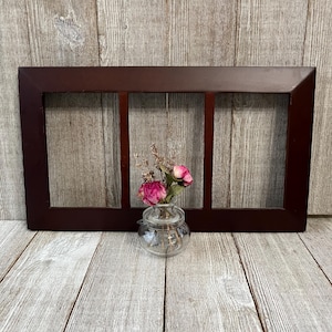 May include: A dark brown wooden frame with three rectangular openings. A small glass vase with pink flowers sits in front of the frame. The background is a wooden surface.