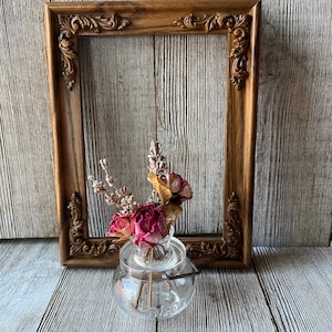 May include: A rectangular wooden picture frame with ornate carved corner details in a medium brown finish, positioned behind a small, clear glass jar containing a bouquet of dried pink roses and sprigs of lavender stems.