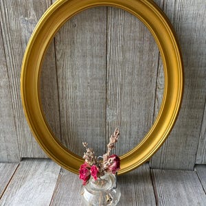 May include: A large, oval-shaped picture frame with a smooth, metallic gold finish. Positioned in front of the frame is a small, clear glass bottle containing a dried arrangement of pink rosebuds and sprigs of lavender.