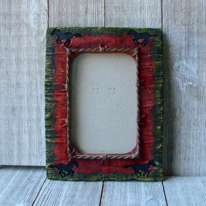 May include: Rectangular picture frame with a rustic, western design. The frame features a red center with black horse silhouettes in each corner. The outer edges are green and the frame has a distressed look.