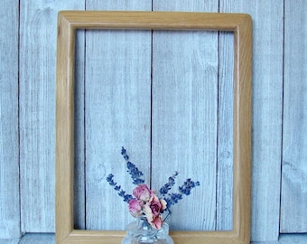 May include: A rectangular, light-brown wooden picture frame with a small glass vase holding dried pink roses and purple lavender. The frame is empty, and the background is a light blue wooden wall.