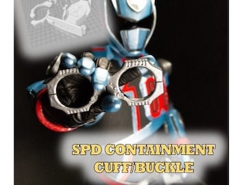 May include: A blue and silver SPD Containment Cuff/Buckle, a prop from the Power Rangers series. The cuff is made of metal and has a textured surface. The buckle is silver and has a smooth surface.