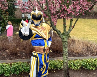 May include: A person in a blue, gold, and white costume with a helmet and a peace sign gesture. The costume has a gold chest plate and belt. The background includes a tree with pink blossoms and a building.