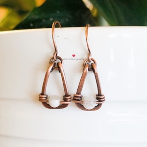 May include: A pair of copper wire earrings with a white moonstone bead in the centre of each earring. The earrings are in the shape of a teardrop.