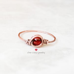 May include: A simple copper wire ring with a red gemstone in the centre. The ring is wrapped in copper wire and has a minimalist design.