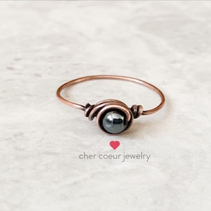 Peut inclure: Une bague simple en fil de cuivre avec une pierre d'hématite noire. La bague est faite à la main et présente un design délicat en fil.