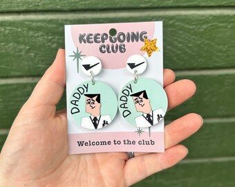 May include: A pair of green and white earrings with a cartoon illustration of a person wearing a suit and the word "Daddy" on each earring. The earrings are on a card that says "Keep Going Club" and "Welcome to the club".