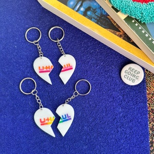 Può includere: Due set di portachiavi a forma di cuore con la scritta "U-Haul" stampata su di essi. I portachiavi sono bianchi con lettere arcobaleno. I portachiavi sono su una superficie blu.
