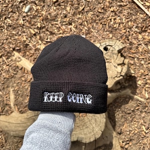 May include: Black knit beanie with white embroidered text that reads "KEEP GOING".