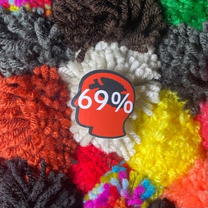 May include: A red and white sticker with a black silhouette of a head and the text "69%" inside. The sticker is placed on a colorful background of fluffy yarn pom-poms.