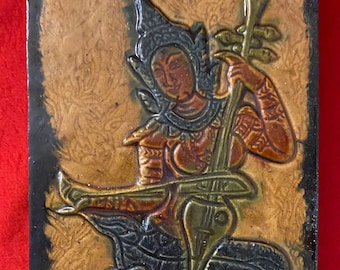 May include: A ceramic tile with a raised relief design of a woman playing a stringed instrument. The woman is wearing a headdress and traditional clothing. The tile is decorated with brown, green, and blue glaze.