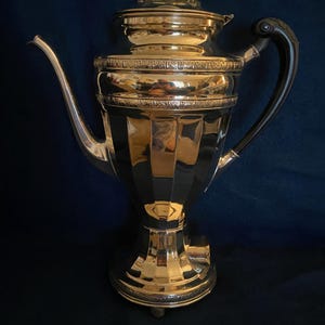 May include: A silver electric coffee pot with a black handle and a decorative design. The pot has a round body with a flared base and a lid with a knob on top.