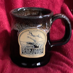 Peut inclure: Une tasse en céramique noire avec un glaçage brun et un autocollant ovale beige. L'autocollant représente un dessin d'un canyon et le texte "est. 1933 Black Canyon of the Gunnison National Park Colorado".