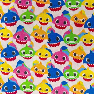 May include: A white fabric with a repeating pattern of colorful cartoon baby shark faces. The sharks have different expressions, including smiling, winking, and wearing glasses.