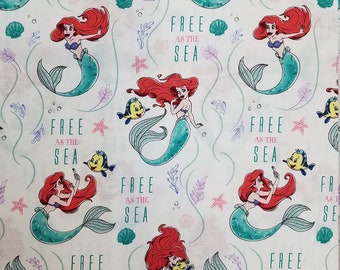May include: A white fabric with a repeating pattern of a red-haired mermaid with a green tail, a yellow fish, and the text "Free as the Sea".