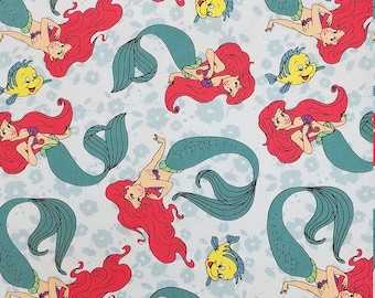 May include: A blue and white fabric with a repeating pattern of red-haired mermaids with teal tails and yellow fish. The mermaids are holding seashells and are swimming in a light blue sea.