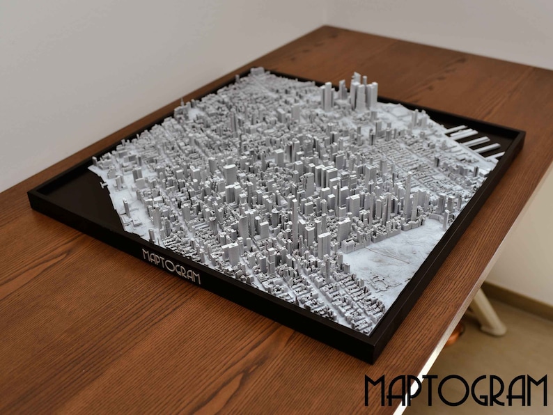 May include: A 3D printed model of a city skyline, with buildings and streets rendered in a silver color. The model is framed in black and sits on a wooden surface. The word "MAPTOGRAM" is printed on the frame.