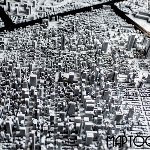 May include: A detailed 3D model of a city skyline, with buildings rendered in shades of gray. The model is set on a black surface and features the word "MAPTOGRAM" in the lower right corner.