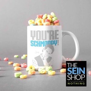May include: White ceramic coffee mug with a gray and blue graphic of Jerry Seinfeld from the TV show Seinfeld. The mug says "You're Schmoopy!" with the word "Schmoopy" in blue. The mug is filled with colorful cereal.