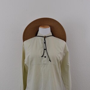May include: A long-sleeved, cream-colored shirt featuring a partial button-down placket, a high collar, and a thin black cord tie with a small wooden bead. The shirt is paired with a wide-brimmed, textured brown straw hat worn on a mannequin.