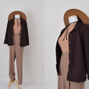 Op de afbeelding: Een bruin knoopshirt met lange mouwen en een kraag. Het shirt wordt gedragen over een lichtbruin tanktop en een bruine broek. Een brede strohoed zit op het hoofd van de mannequin.