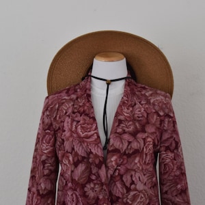 May include: A blazer featuring a dark pink and maroon floral pattern, worn over a white shirt. A wide-brimmed brown straw hat is positioned behind the collar, and a thin black cord necklace with a small brown bead hangs around the neck area.