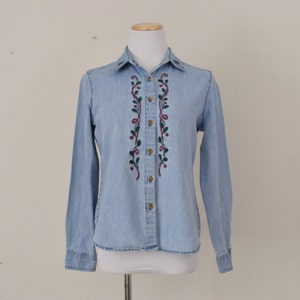 Puede incluir: Una camisa vaquera azul claro de manga larga y cierre de botones. La camisa presenta un diseño bordado de enredaderas verdes con bayas rojas y una cinta dorada.