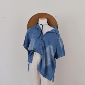 May include: A denim button-up shirt with short sleeves and a faded wash. The shirt has a collar and is adorned with small, scattered white embellishments. It is paired with light-colored pants and a wide-brimmed brown hat.