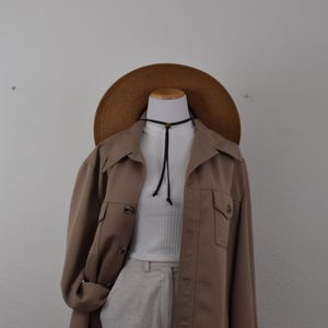 May include: A tan long-sleeved button-up jacket worn over a white ribbed mock-neck top, paired with light-colored trousers. A wide-brimmed brown straw hat sits above the neckline, and a thin black cord necklace with a small bead is visible.