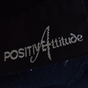 Pu&ograve; includere: Un'etichetta di abbigliamento nera con testo bianco che recita "POSITIVE ATTITUDE" in un carattere stilizzato.