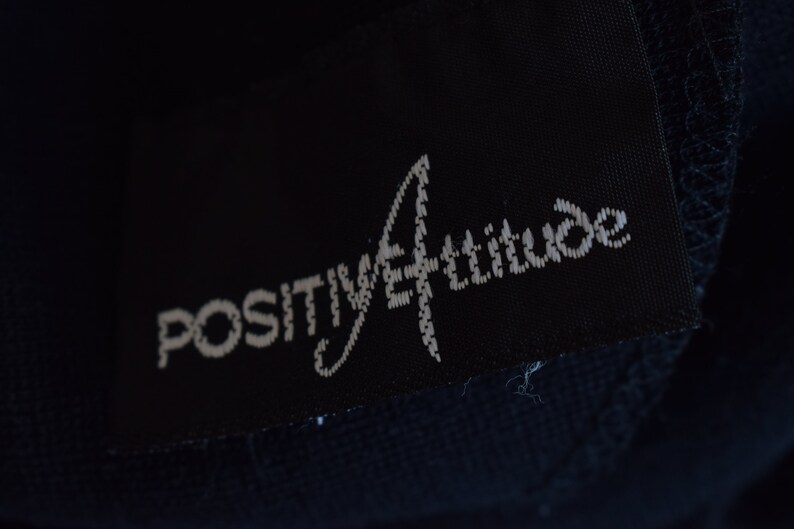 Pu&ograve; includere: Un'etichetta di abbigliamento nera con testo bianco che recita "POSITIVE ATTITUDE" in un carattere stilizzato.