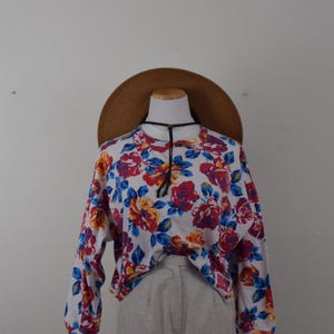 May include: A long-sleeve white sweatshirt featuring a vibrant floral pattern in shades of red, orange, purple, and blue. The garment includes a black drawstring cord at the neckline and is paired with a wide-brimmed brown straw hat.