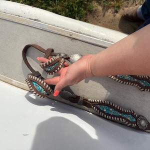 May include: Brown leather horse tack with turquoise and silver accents. The tack features decorative stitching and silver conchos. The design includes a turquoise center with silver studs and a white border. The leather is a rich brown color.