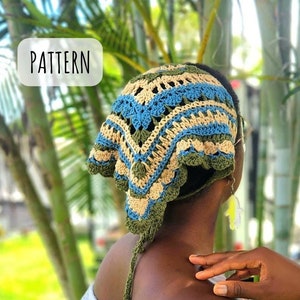 Op de afbeelding: Een vrouw draagt een gehaakt hoofddoek met een groen, blauw en beige patroon. De doek is vastgebonden aan de achterkant van haar nek en heeft een lange kwast. Ze draagt een witte kanten jurk.