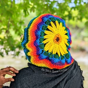 May include: A colorful crocheted beanie with a sunflower design. The beanie is made with a rainbow of colors, including red, orange, yellow, green, blue, and purple. The sunflower is in the center of the beanie and has a brown center and yellow petals. The beanie is worn by a person with black clothing. The text "The Stitch Queen Crochet" is visible on the beanie.