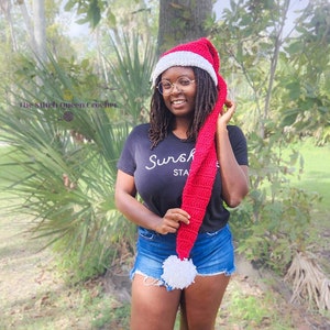 May include: A woman wearing a red and white crocheted Santa hat with a long, dangling tassel. She is wearing a black t-shirt with the text "Sunshine State" and denim shorts.