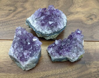 May include: Three amethyst crystal clusters with deep purple crystalline structures. The crystals are set on a grey and white base, resting on a wooden surface. The clusters vary in size and shape, showcasing natural formations.